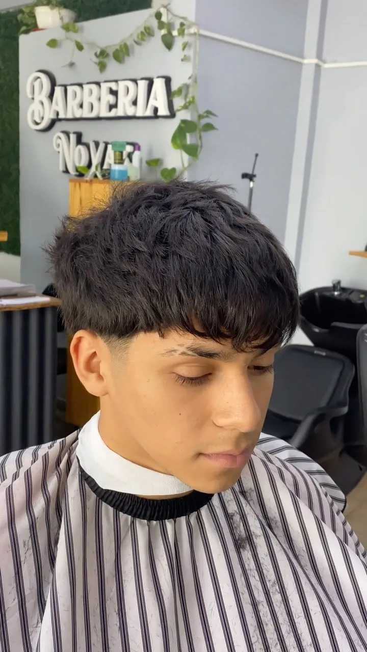 Hombre joven de frente en la barbería con taper fade, fleco texturizado y una raya fina en la ceja derecha