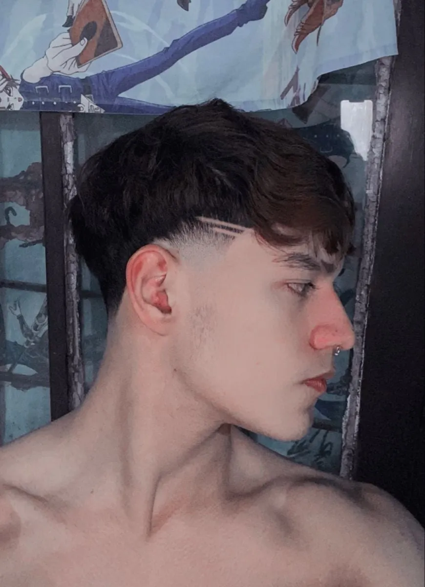 Taper fade de perfil con dos rayitas finas en la patilla sobre la oreja y cabello castaño oscuro con fleco largo