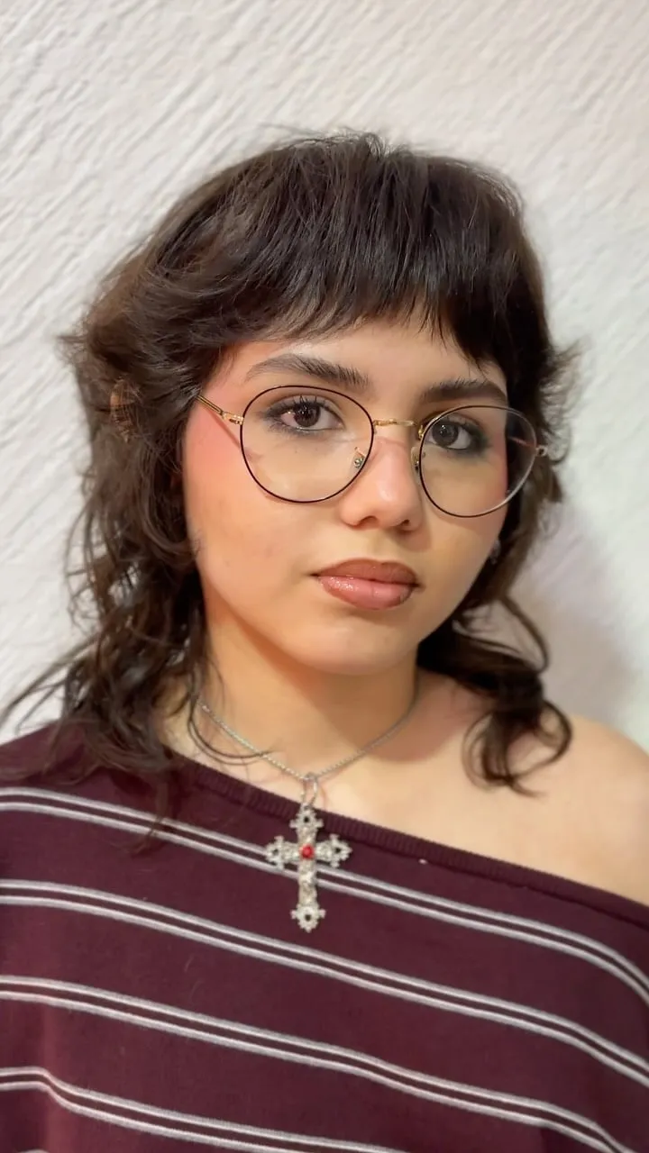 Mujer joven con lentes y wolf cut castaño oscuro, flequillo corto por encima de las cejas y capas desfiladas a la altura del cuello