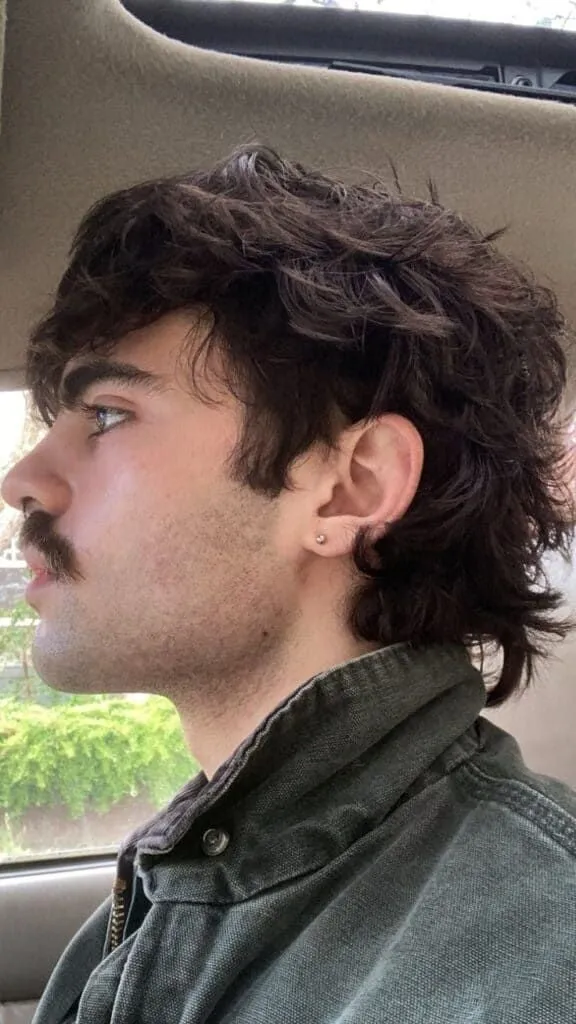 Hombre de perfil con bigote y wolf cut en cabello castaño oscuro ondulado, volumen arriba y nuca desfilada al cuello