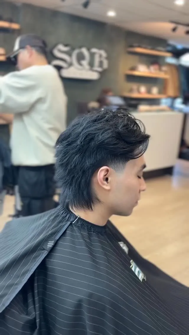 Joven de perfil en la barbería con wolf cut negro, mid fade alrededor de la oreja y nuca larga en punta