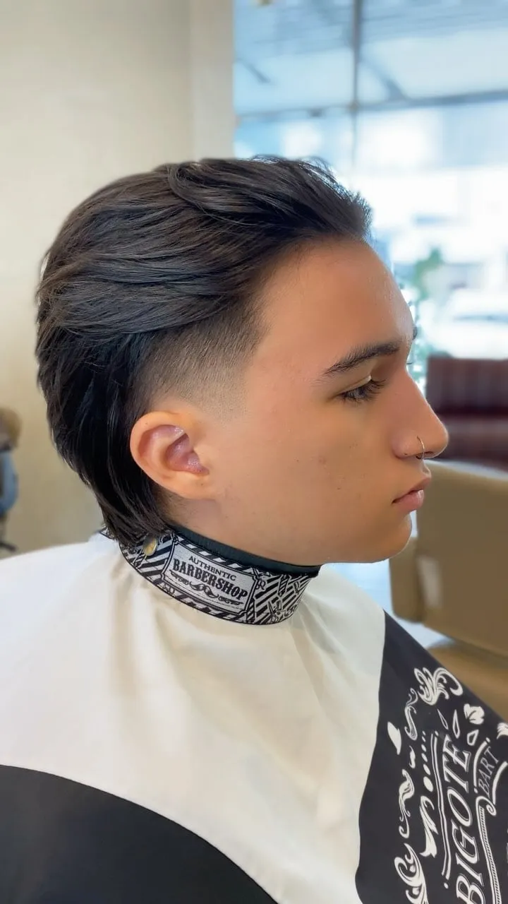 Joven de perfil en la barbería con wolf cut castaño oscuro, taper fade en la patilla y nuca larga desfilada